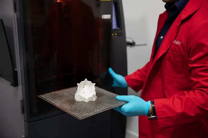 Forge Labs technician holding a white SLA part on its build plate beside an SLA printer