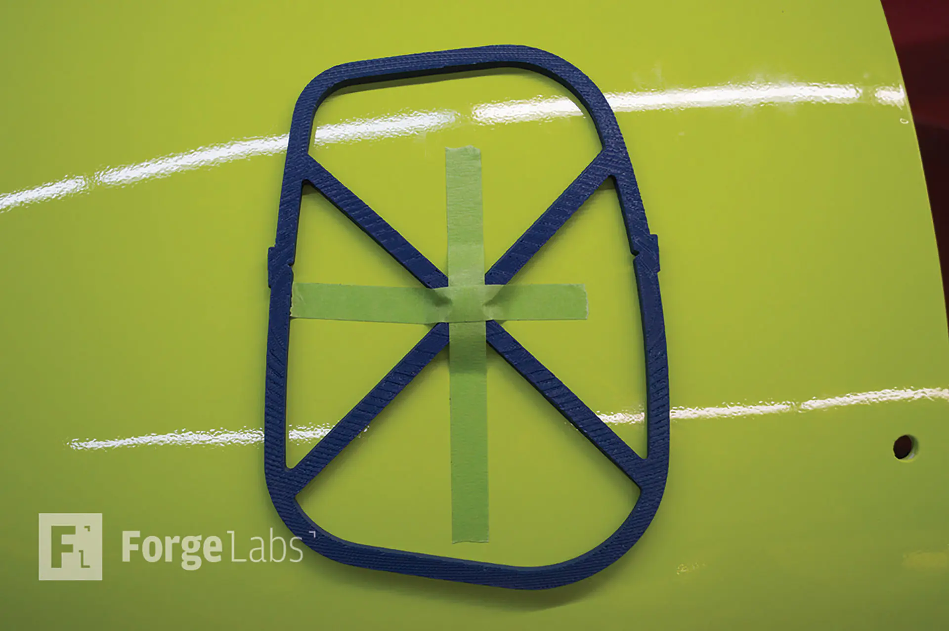 A blue FDM-printed open-frame template with diagonal braces taped to the glossy yellow-green curved hull of an underwater vehicle
