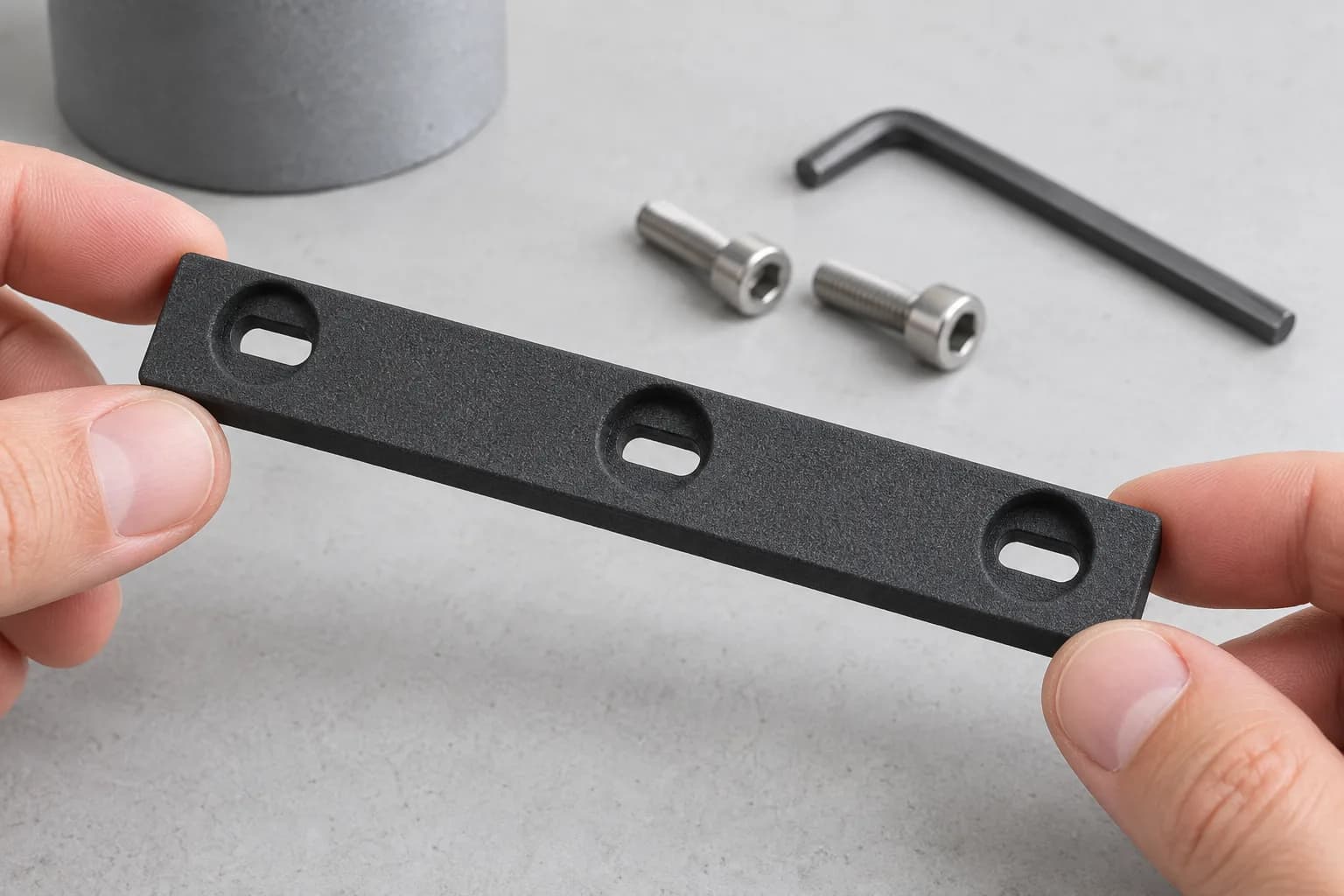 Fingertips holding a flat matte black SLS nylon mounting strip with slotted, counterbored holes above a clean bench